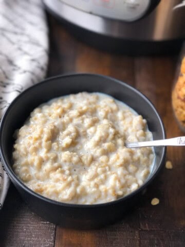 A black bowl of oatmeal with a silver spoon in the bowl on a wooden table with a towel to the left of the bowl.