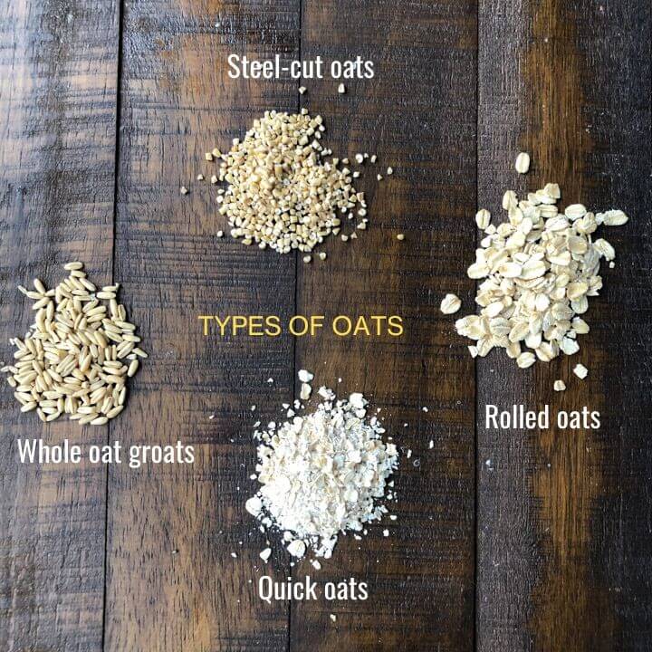 4 piles of oats on a wooden table. The pile at the top shows steel-cut oats. To the right is a pile of rolled oats. The bottom pile labeled quick oats, and the pile to the left labeled whole oat groats.