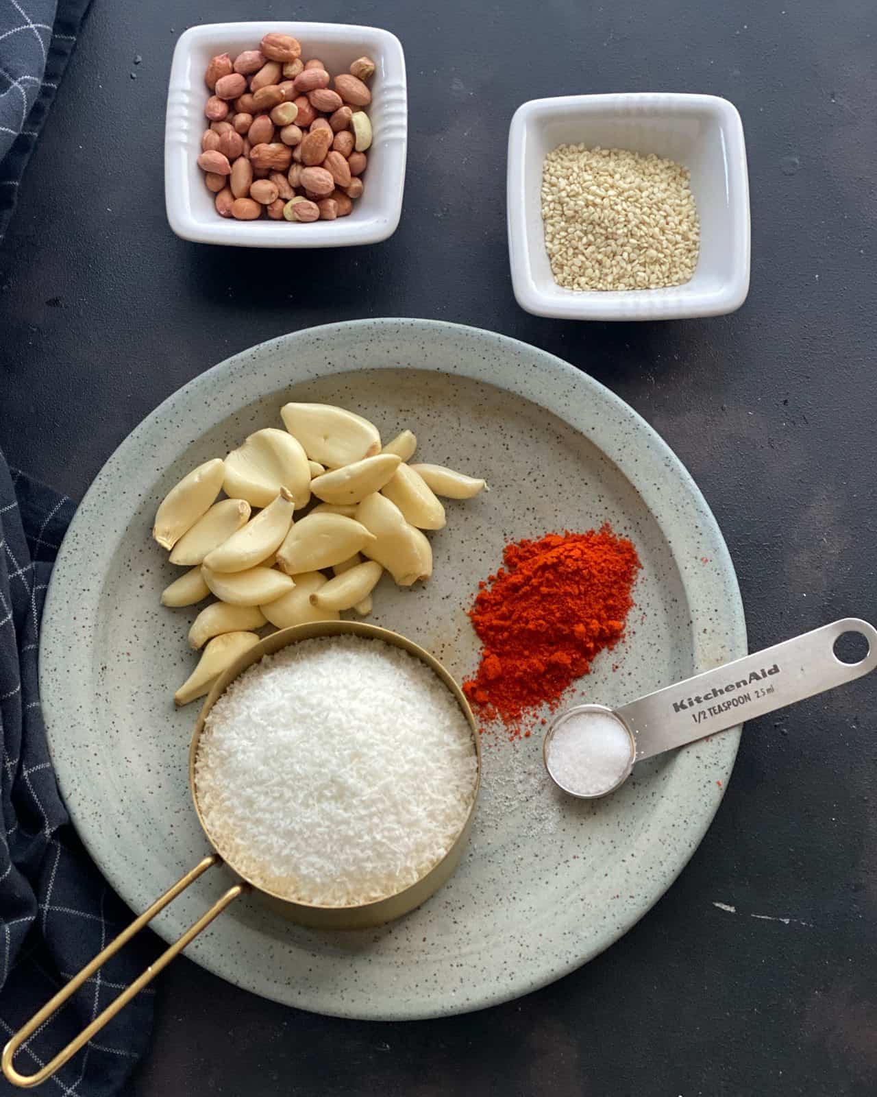 Ingredients for garlic chutney laid out in grey plate - desiccated coconut, chili powder, garlic cloves, salt. Sesame seeds and red skin peanuts are in separate bowls.