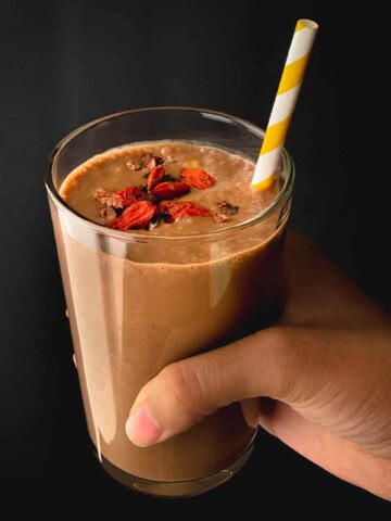 A hand holding a chocolate date smoothie in a clear glass cup with goji berries on the top of the smoothie and a yellow and white straw in the smoothie.