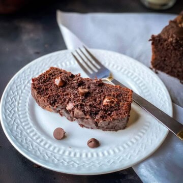 A small white plate with a slice of chocolate zucchini bread and a silver fork next to the slice of moist double chocolate bread.