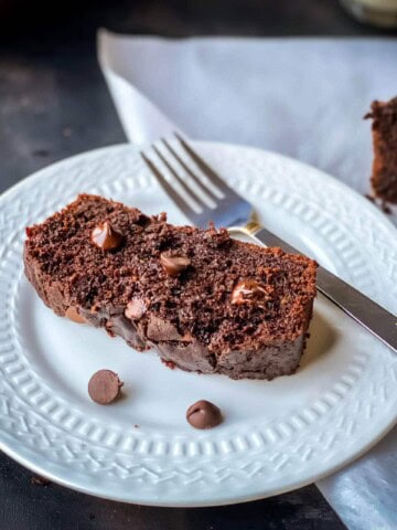 A small white plate with a slice of chocolate zucchini bread and a silver fork next to the slice of moist double chocolate bread.