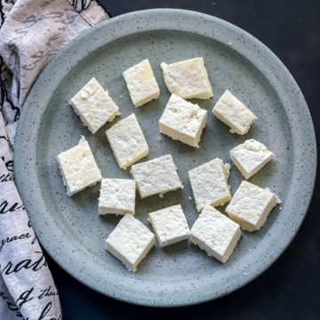 Cut cubes of paneer on a light blue speckled plate with a dish towel to the left.