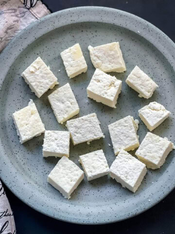A blue speckled plate with cubes of instant pot homemade paneer with a dish towel on the left.