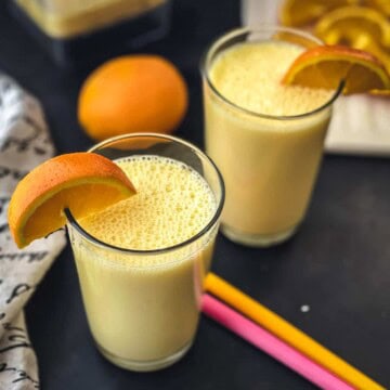 Two glasses of orange smoothie with wedges of orange on the rim, two colorful straws in the middle of the smoothie cups, and an orange in the back.