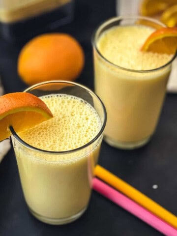 Two glasses of orange smoothie with wedges of orange on the rim, two colorful straws in the middle of the smoothie cups, and an orange in the back.