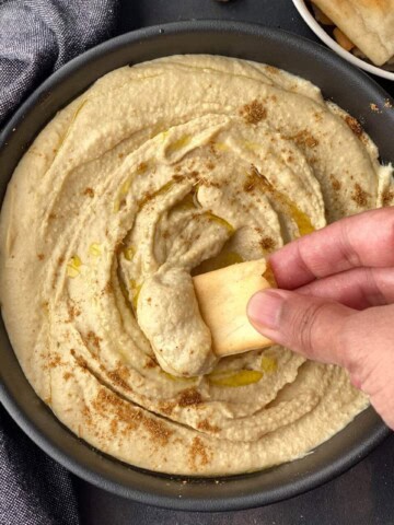 A hand dipping a pita chip into a black bowl filled with creamy instant pot hummus.