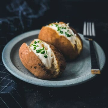 A black counter with a blue plate and two baked potatoes cut down the center vertically and topped with sour cream and chives with a silver fork on the right side of the plate.