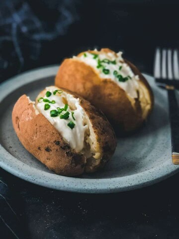A black counter with a blue plate and two baked potatoes cut down the center vertically and topped with sour cream and chives with a silver fork on the right side of the plate.