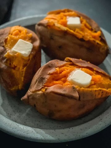 A light blue plate with three baked sweet potatoes with a cut down the middle of each and a pad of butter on top.