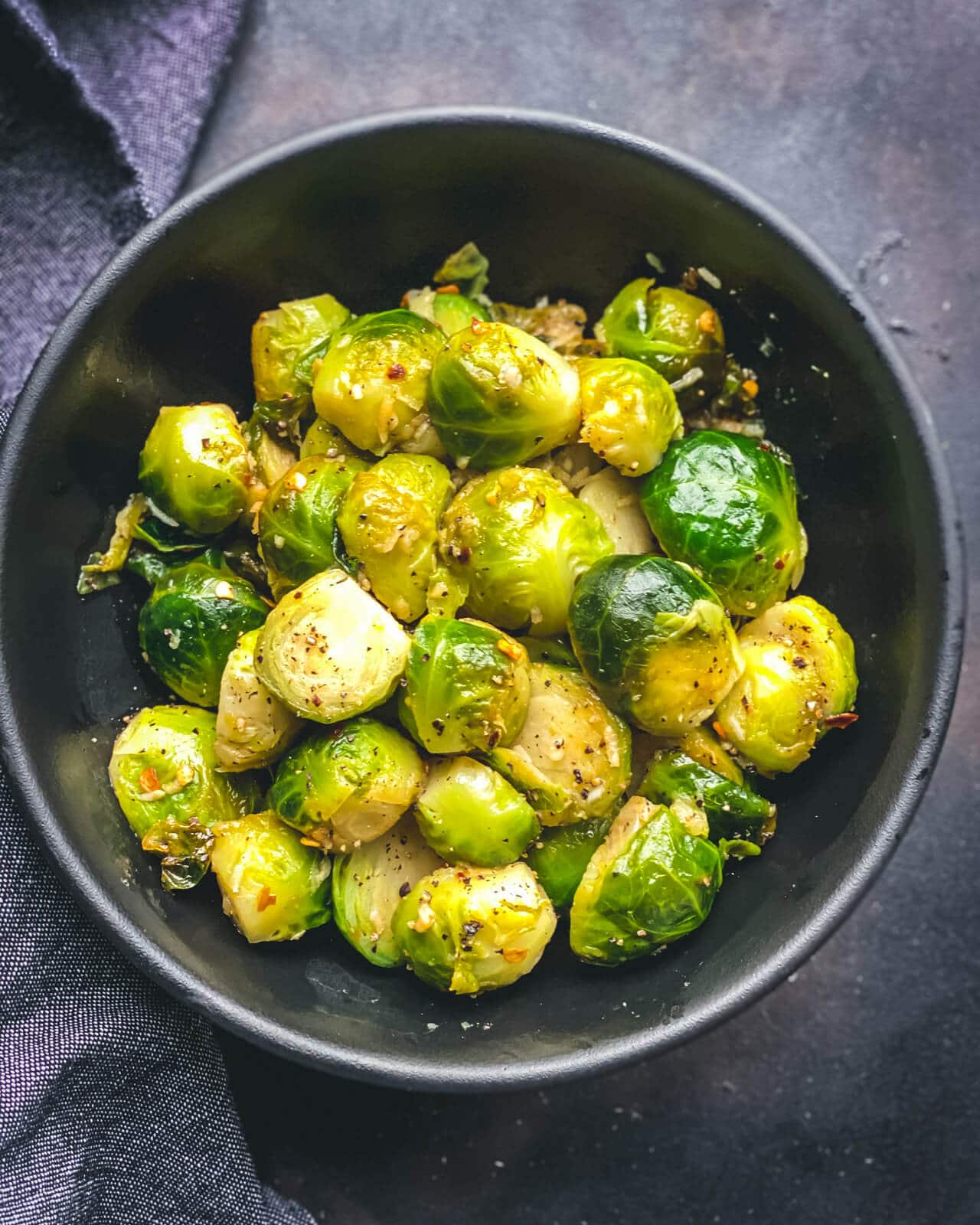 A black bowl with bright green steamed Brussel Sprouts on a grey counter with a grey blue towel to the left.
