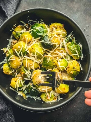 A black bowl with steamed and sautéed Brussel sprouts topped with cheese and a silver fork being poked into the bowl on a grey counter.