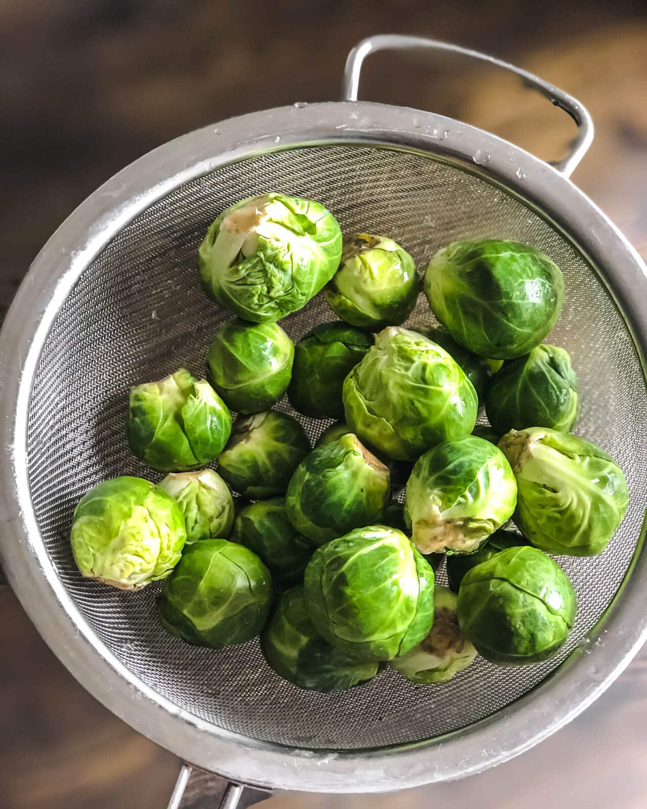 A silver mesh strainer with brussel sprouts over a wooden counter.