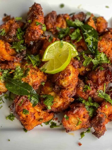 A white plate with an array of crispy chicken 65 topped with cilantro and lemon.