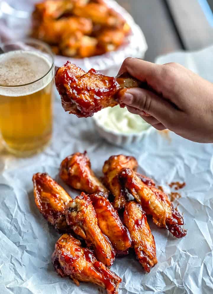 A hand holding a bbq chicken wing over a stack of other wings with a cup of ranch and a glass of beer in the back.
