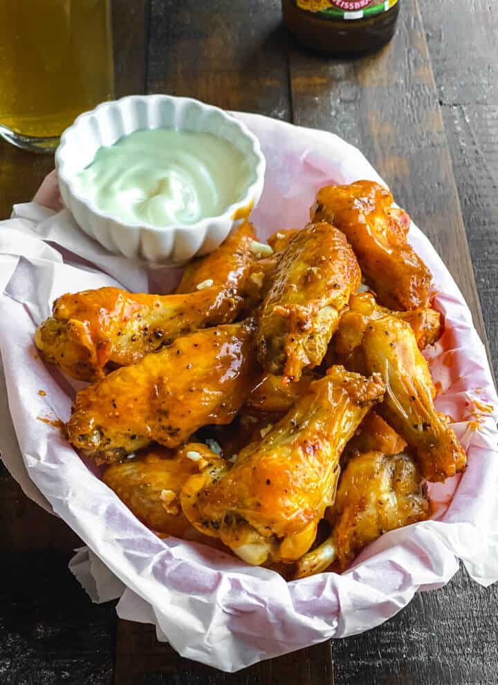 A parchment paper lined basket with a stack of buffalo chicken wings with a small cup of ranch in the back.