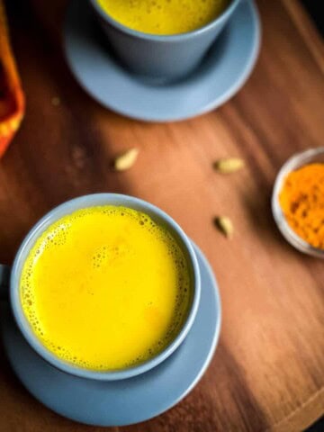 A blue mug with golden turmeric latte on a wooden counter with another latte in the back and a small bowl of turmeric to the right.