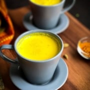 A pair of grey-blue mugs on a wooden counter filled to the brim with a bright golden mixture of turmeric milk with the words Step by Step Golden Turmeric Milk at the bottom.