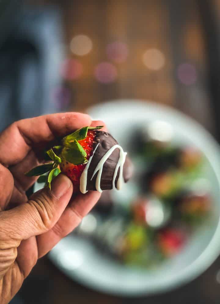 A hand holding a chocolate covered strawberry drizzled with white chocolate in front of a plate of chocolate covered strawberries.