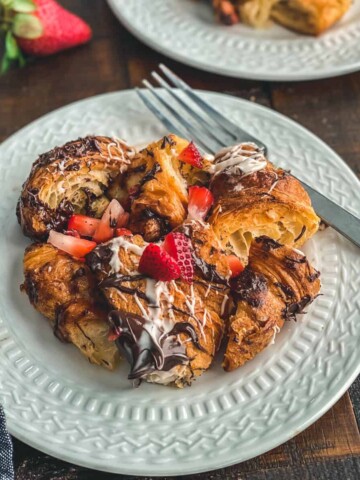 A white plate with a serving of chocolate croissant bread pudding topped with melted chocolate and fresh fruit.
