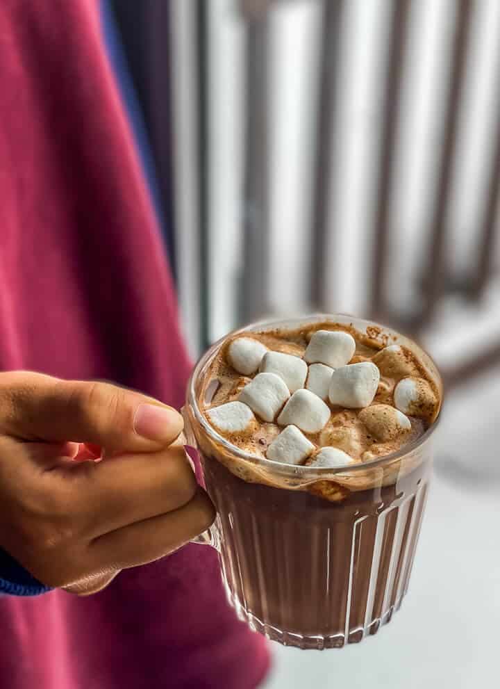 A hand holding a glass mug of hot chocolate topped with marshmallows.