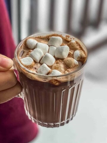 A square picture with a hand holding a clear glass mug of hot chocolate topped with marshmallows.
