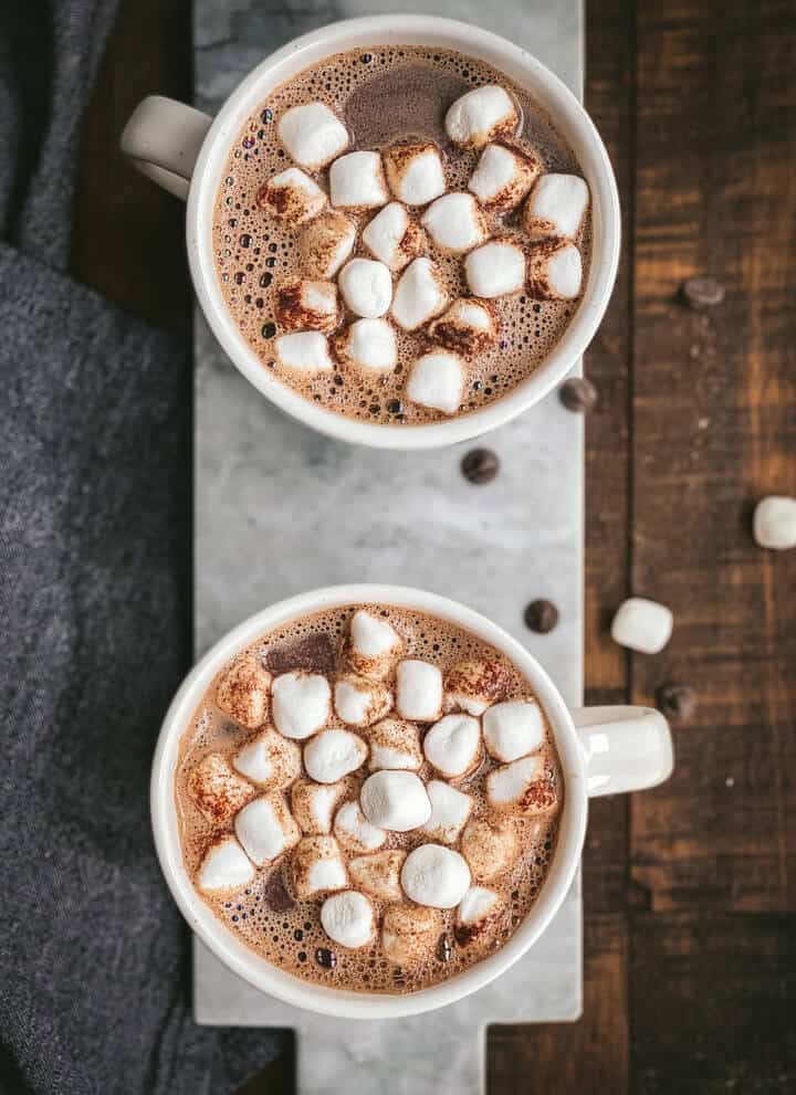 Two white mugs of hot chocolate topped with marshmallows on a marble cutting board.