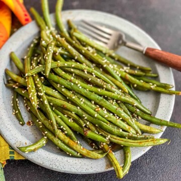 A light blue plate with a pile of asian-style green beans topped with toasted sesame seeds and a brown handled fork in the back right.