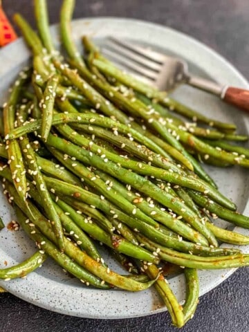 A light blue plate with a pile of asian-style green beans topped with toasted sesame seeds and a brown handled fork in the back right.
