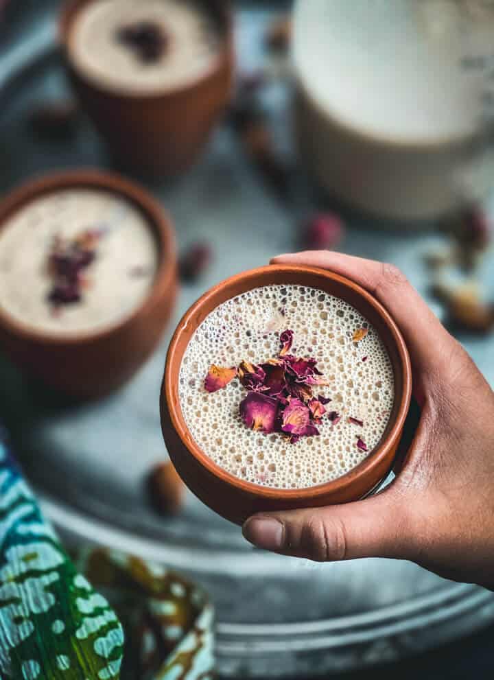 A hand holding a brown mug with thandai and topped with rose petals in front of a tray of thandai. 