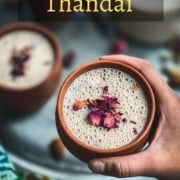 A hand holding a brown mug with thandai topped with rose petals in front of a silver plate with other mugs of thandai topped with rose petals and the word Thandai in yellow in the top middle of the picture.