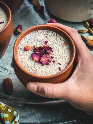 A hand holding a brown mug of thandai topped with rose petals over a silver platter with another mug of thandai to the left.
