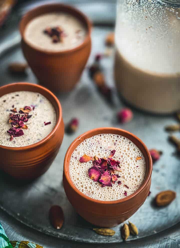 A silver platter with brown mugs of thandai topped with rose petals and a glass mug of thandai in the back right.