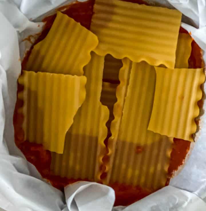 A springform pan lined with parchment paper with sauce and a layer of noodles.