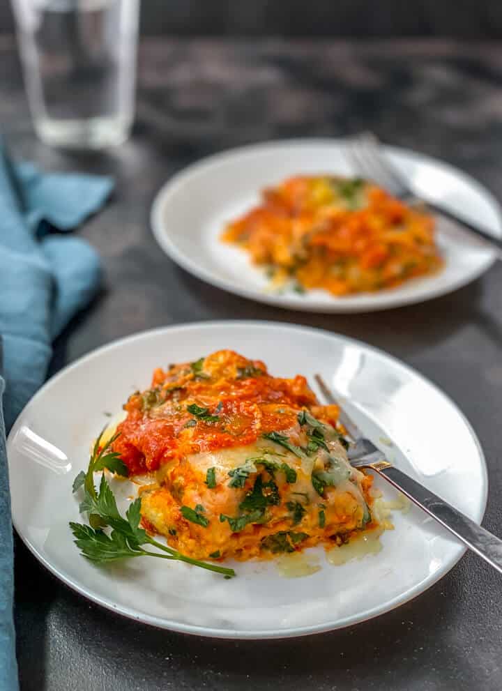 A white plate with a serving of instant pot lasagna with a silver fork and another serving of lasagna on a white plate in the back with a blue towel to the left.