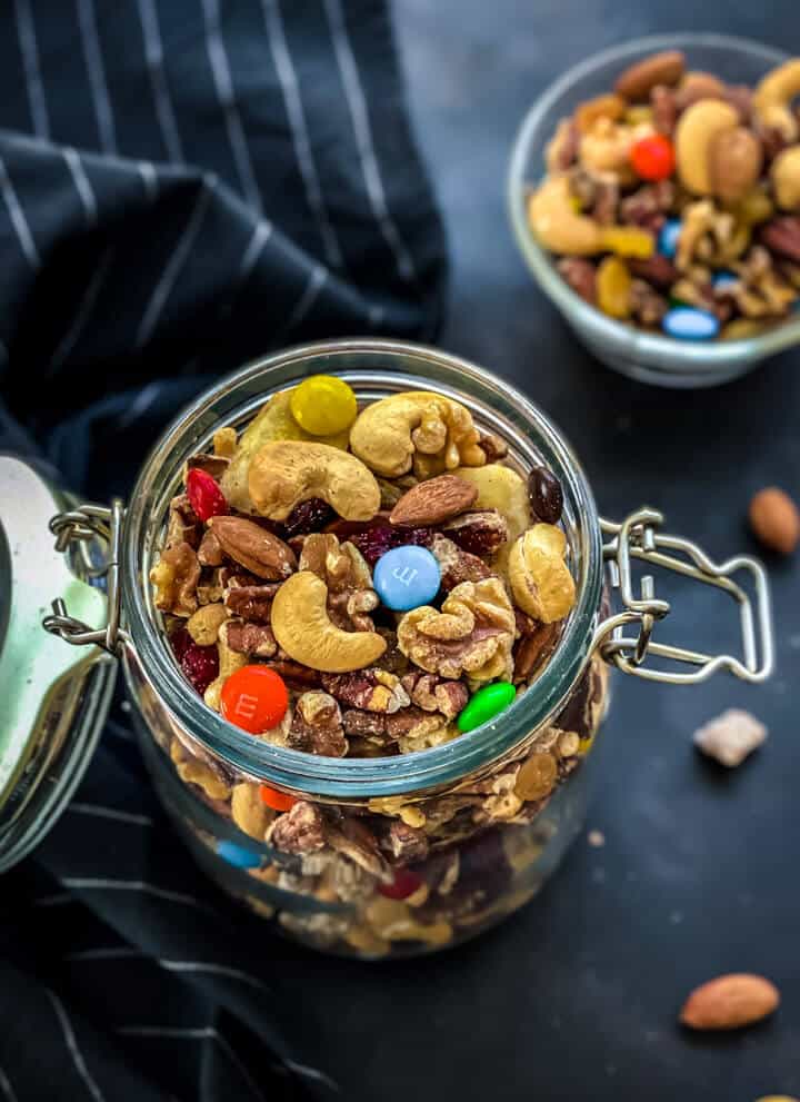 A glass jar filled with easy trail mix with a small bowl of homemade trail mix in the back on a dark grey counter.