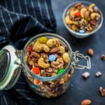 A glass jar with a lid attached on the left side with the lid hanging open and homemade trail mix inside the jar with a small glass bowl of homemade trail mix in the back.