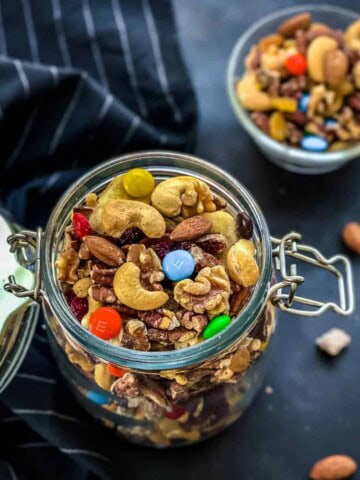 A glass jar with a lid attached on the left side with the lid hanging open and homemade trail mix inside the jar with a small glass bowl of homemade trail mix in the back.