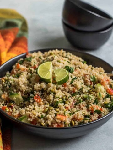 A black bowl with quinoa salad topped with two lime slices and a fork in the right side of the bowl with two small black bowls in the back and a colorful dish towel to the left.