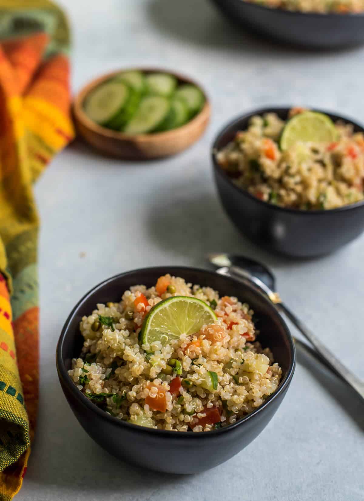 Two small black bowls of quinoa salad with a spoon in between the two bowls and a small brown bowl to the back left with cucumber slices and a colorful dish towel on the left side of the photo.