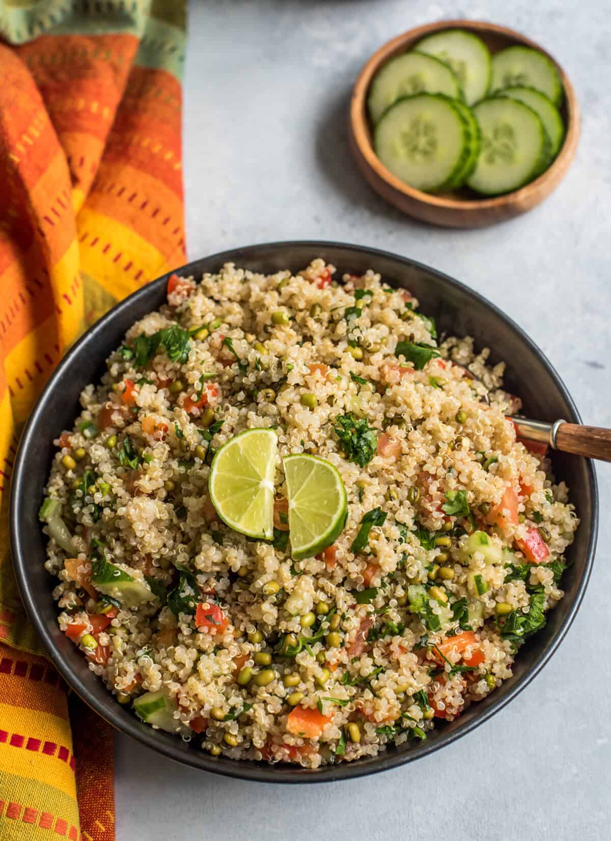 A top down photo of a black bowl filled with easy quinoa salad and topped with two slices of lime and a spoon in the top right of the bowl with a colorful dish towel to the left side and a small wooden bowl with cucumbers in the back.