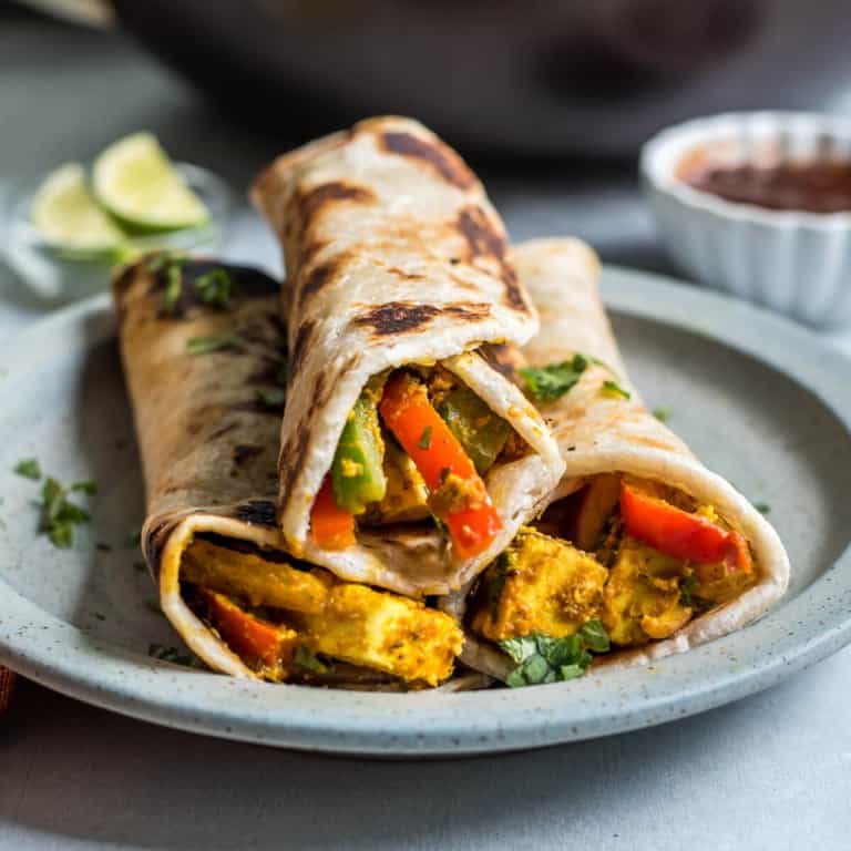 A light blue plate with three paneer kathi rolls stacked in a pyramid with a small white bowl of tamarind chutney in the back right.