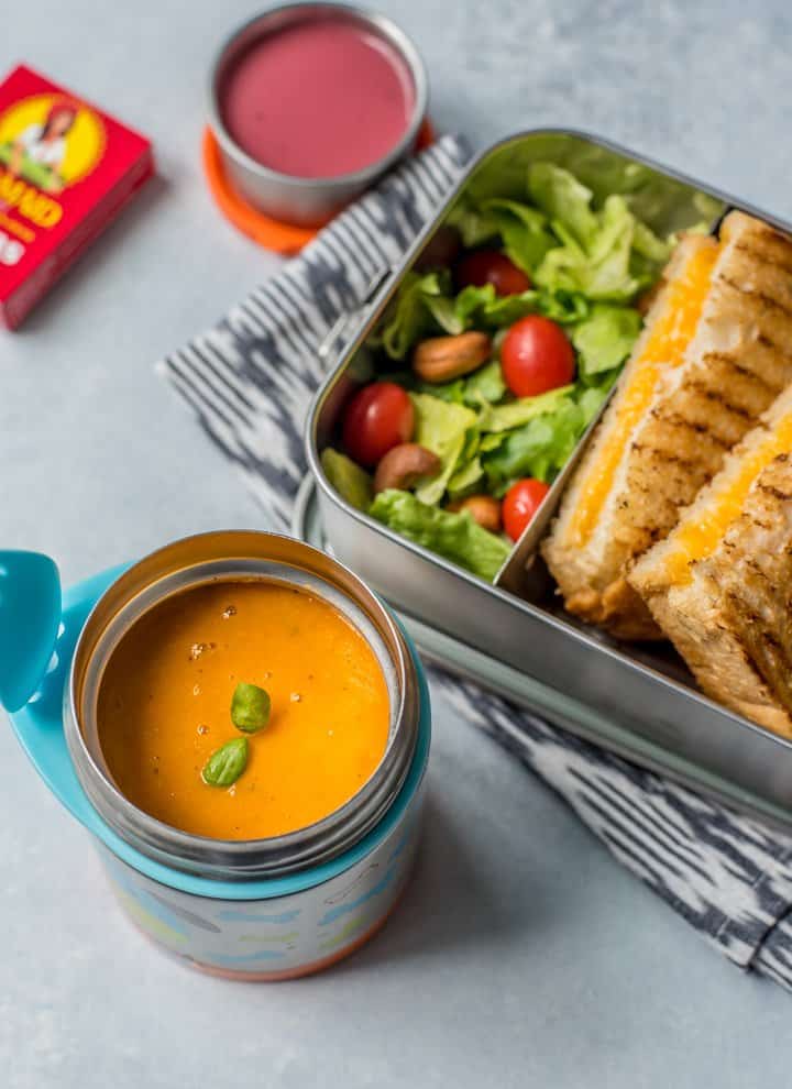 Tomato soup in a thermos with grilled cheese in a steel container along with salad and dressing. Raisins are on the side.