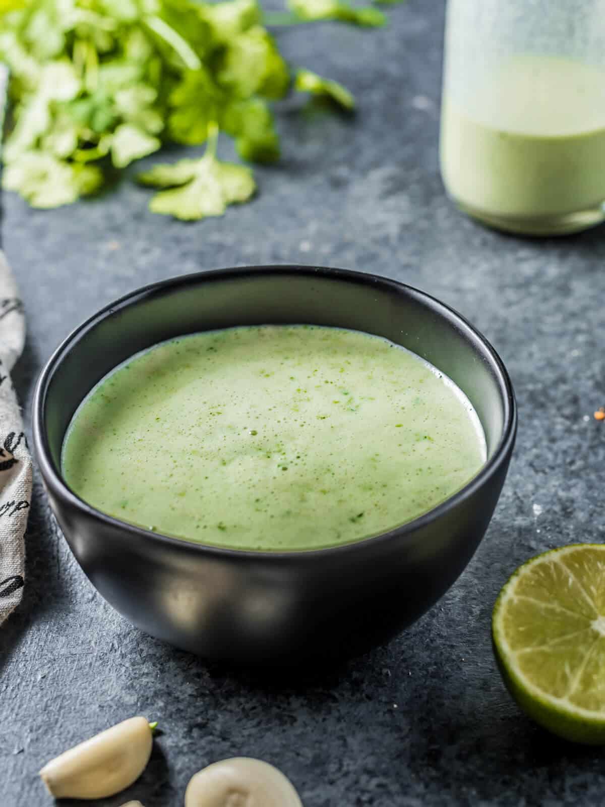 A bowl that contains a creamy green dressing. A bunch of cilantro in the background, with garlic cloves and limes in the foreground. 