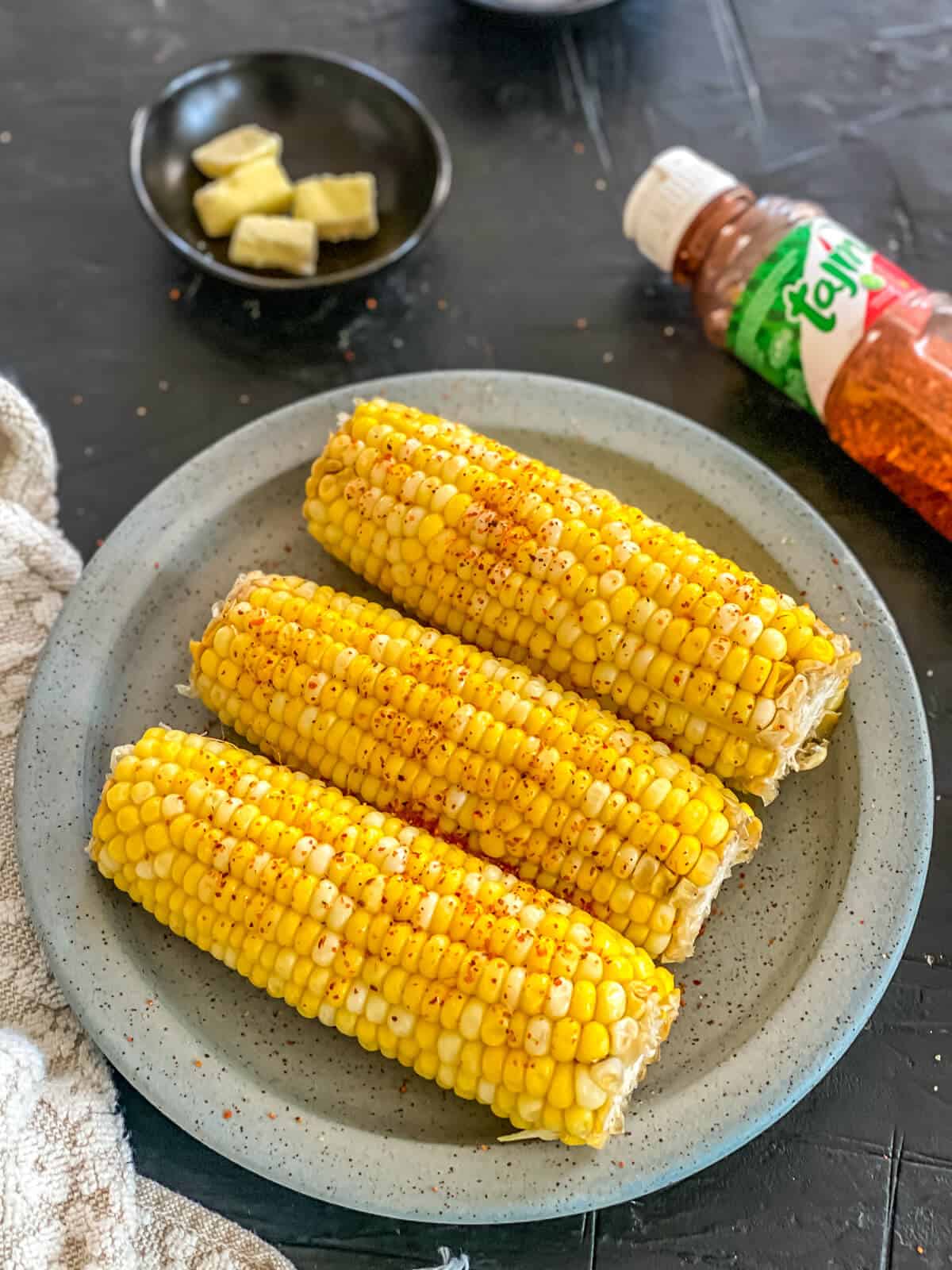 Three cobs of corn, cooked and laying on a plate. Sprinkled with seasoning.