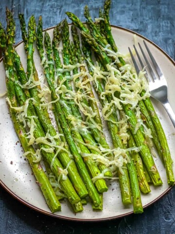 A cream plate on a blue counter with air fried asparagus topped with parmesan and a silver fork to the right.