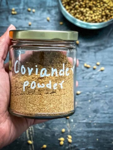 A hand holding a glass jar labeled coriander powder over a grey counter with coriander seeds.