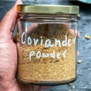 A hand holding a glass jar labeled coriander powder over a grey counter with coriander seeds and the words Make This At Home Coriander Powder at the top.