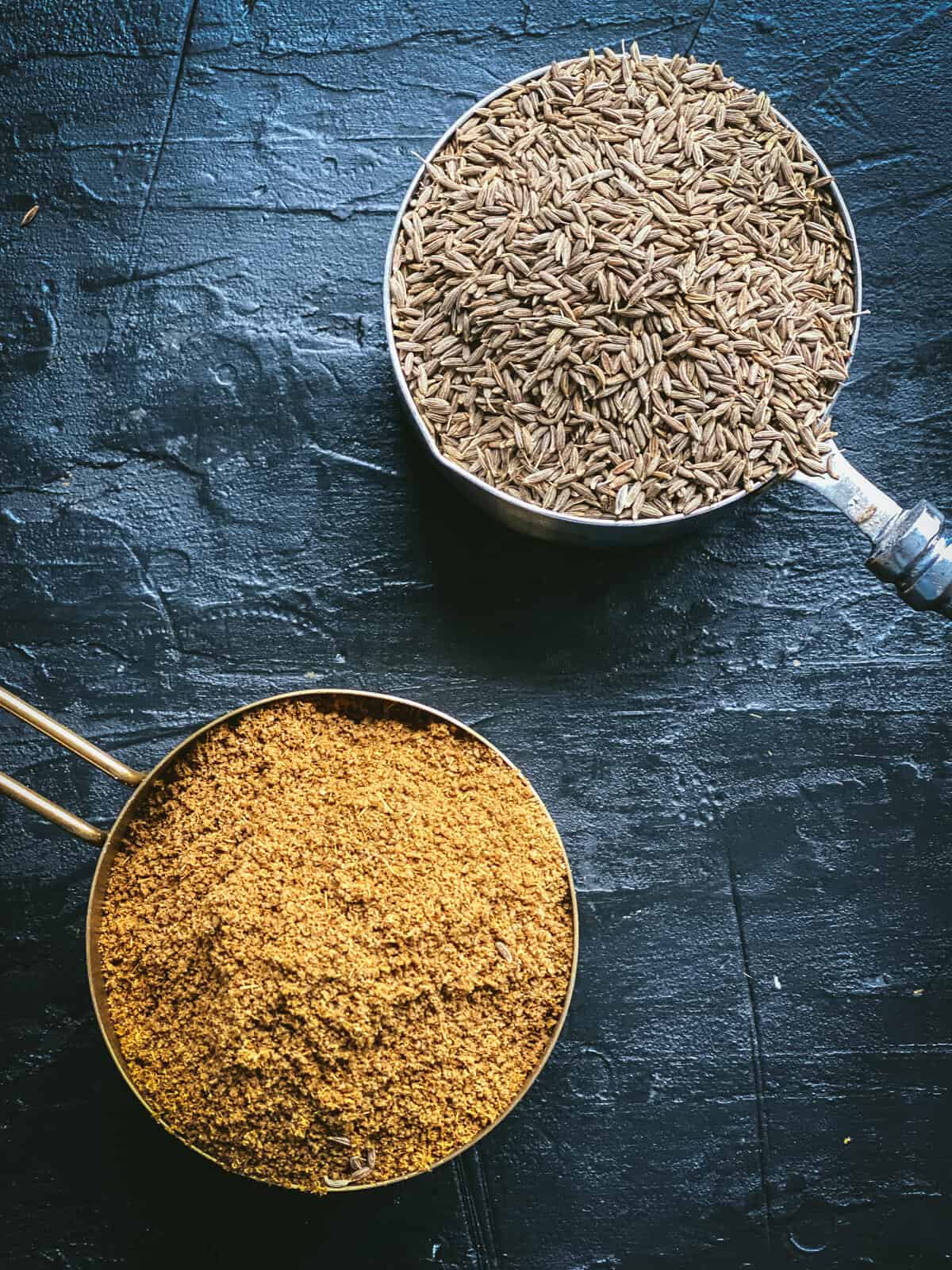 Two measuring cups: one with cumin seeds, and the other with ground cumin powder.