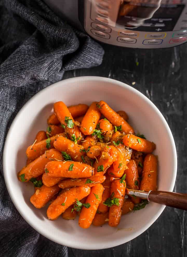 A white bowl with steamed carrots and a fork on the right side of the bowl with an instant pot in the back right.